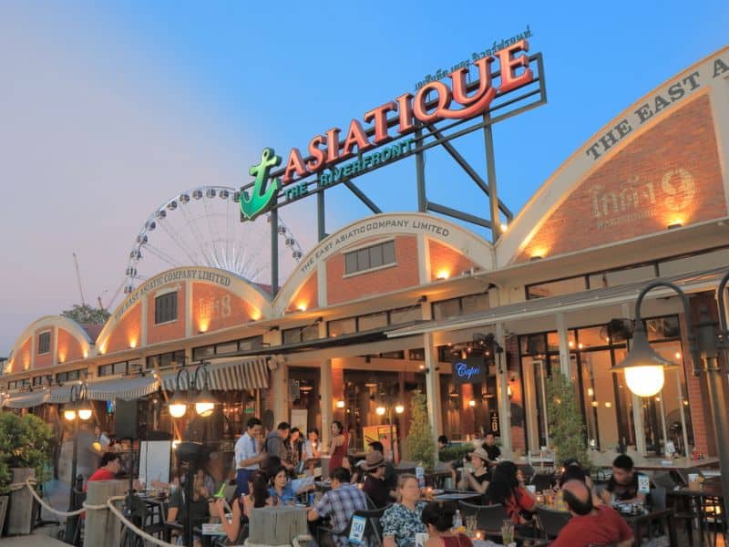 People at an outdoor area at an open-air mall Asiatique in Bangkok, Thailand with a ferris wheel in the background.