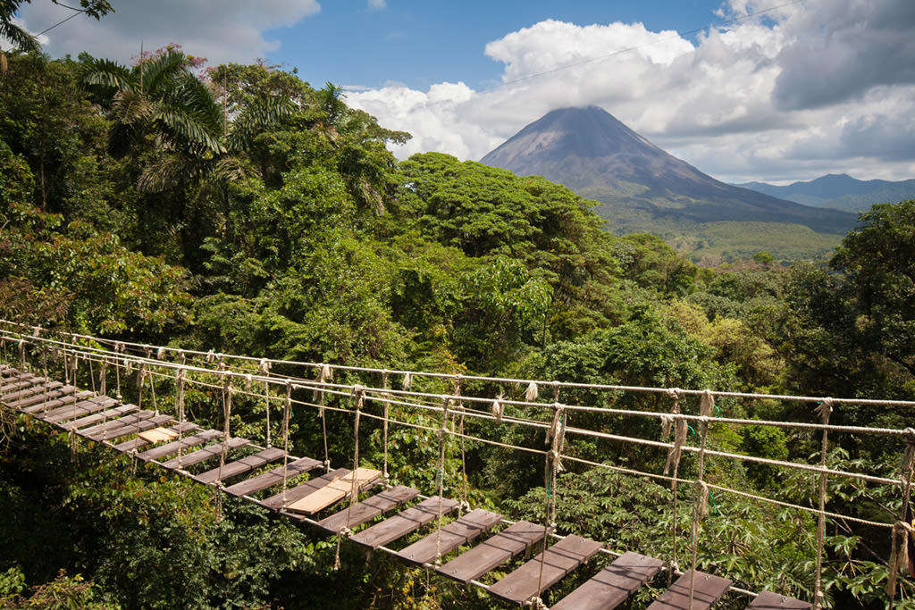 Hanging bridge stretches across lush green canopy in La Fortuna, with Arenal Volcano visible in the distance, in Costa Rica