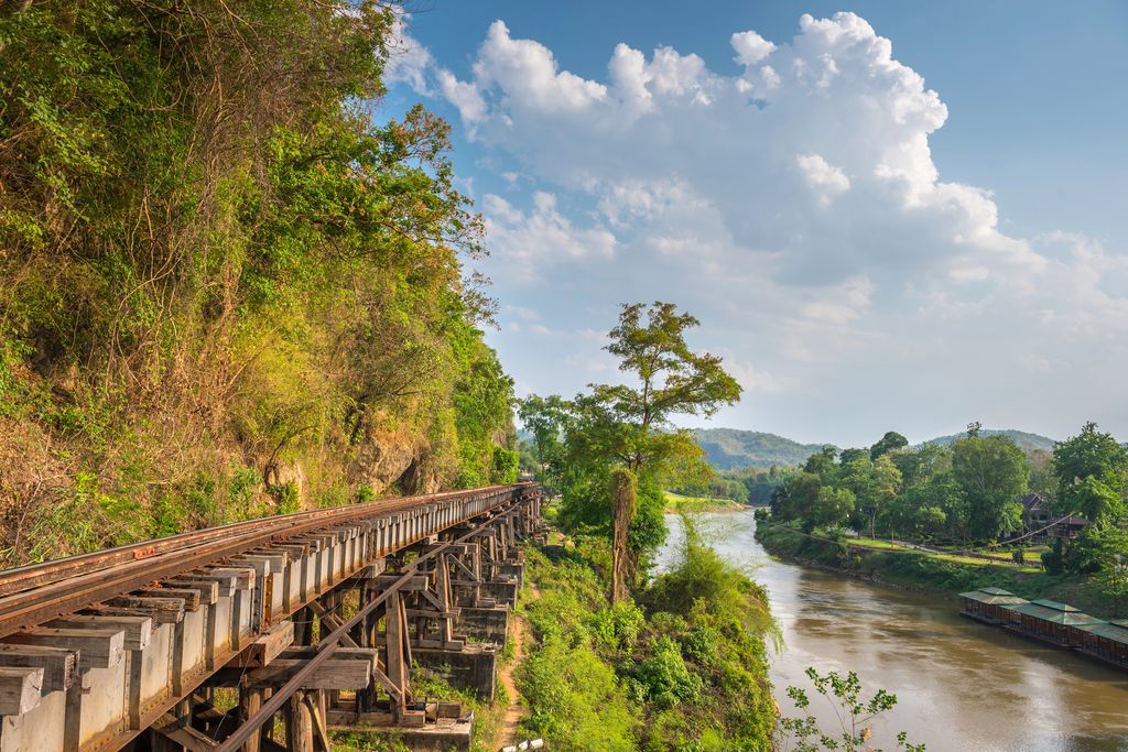 A railroad by the river surrounded by trees on a sunny day.