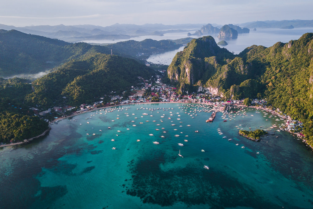 Aerial view of El Nido, Palawan, showing a vibrant coastal town, turquoise waters dotted with boats, and towering limestone cliffs in the background.