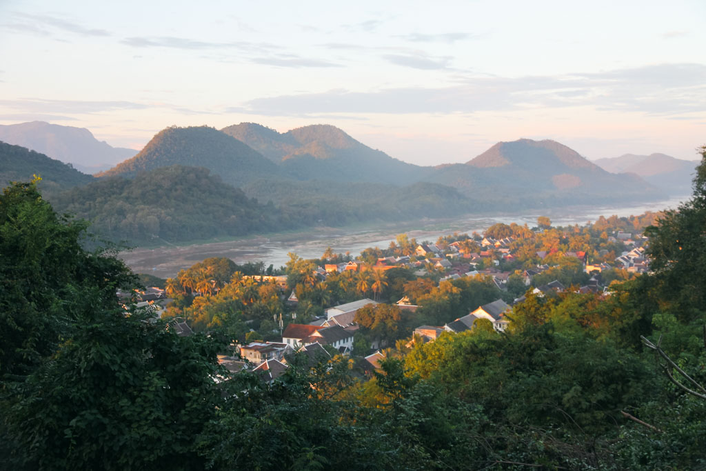 A small town in Laos with houses, trees, and river and hills in the background.