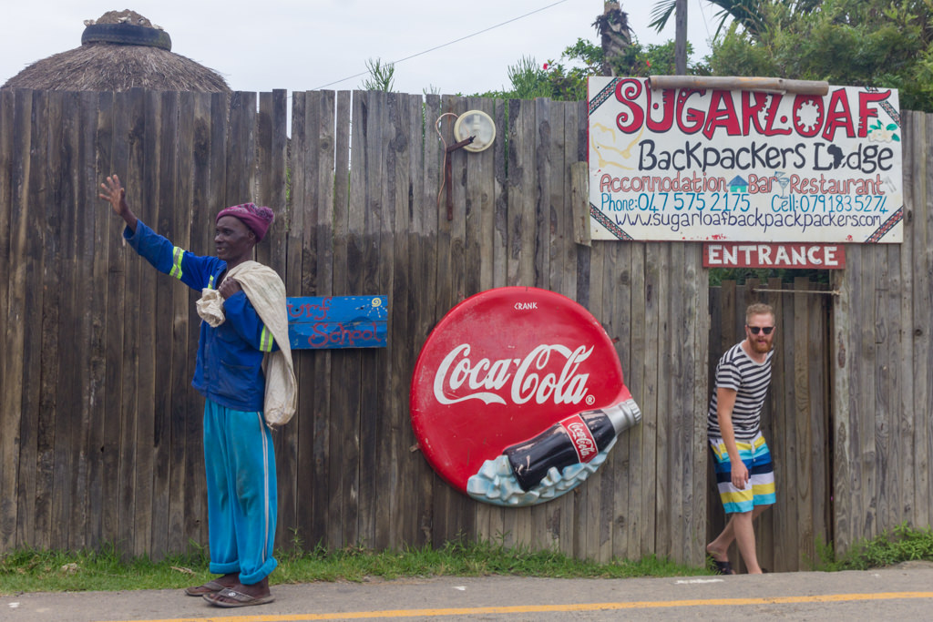 A backpacker exiting a hostel in Coffee Bay while a local hails a bus