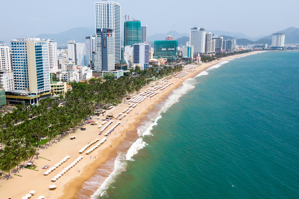 The coastline of a beach on Nha Trang in Vietnam surrounded by the sea, trees, and buildings.