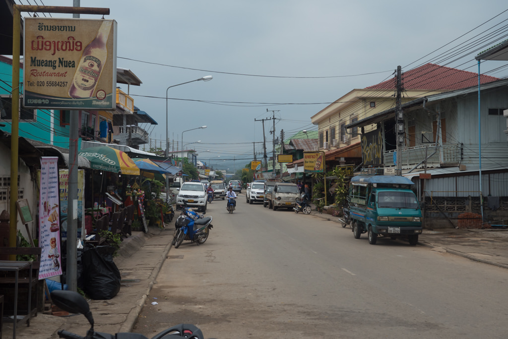 A road in Laos with motorcycles and cars parked on the sides.
