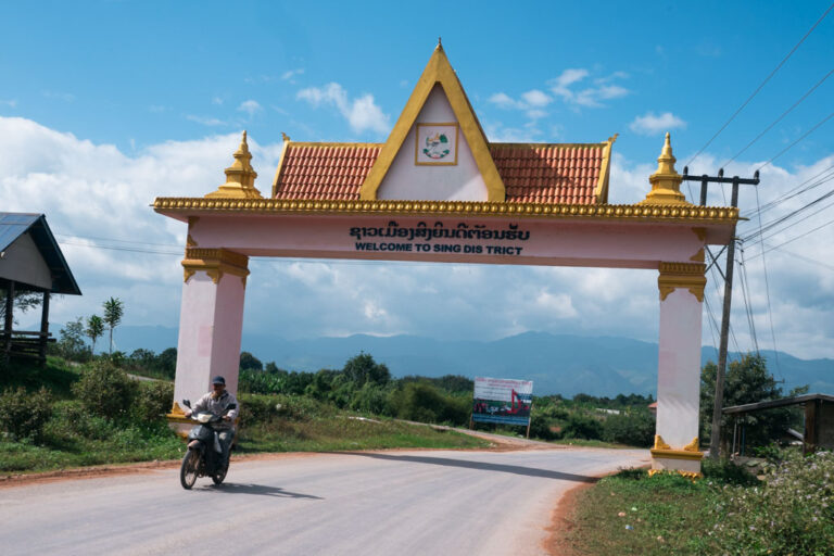 A person riding a motorcycle on a road in Luang Namtha, Laos and an arch in the background.