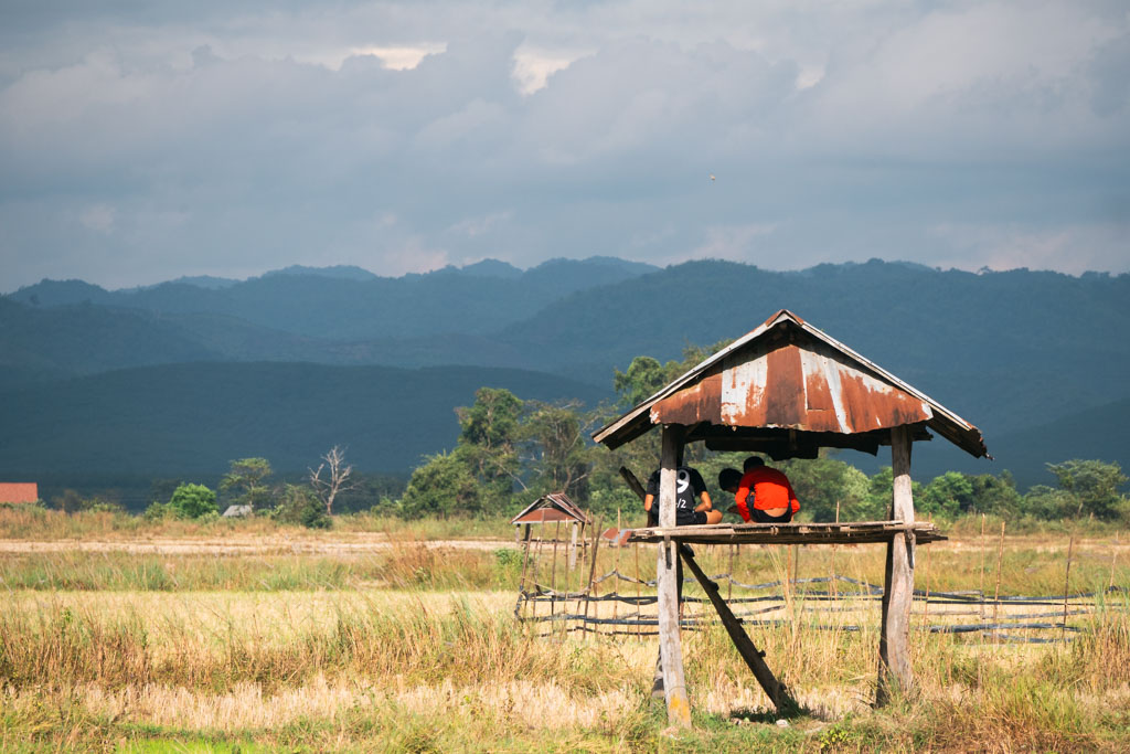 People sitting on a shed at a rice field in Luang Namtha, Laos with mountains in the background.