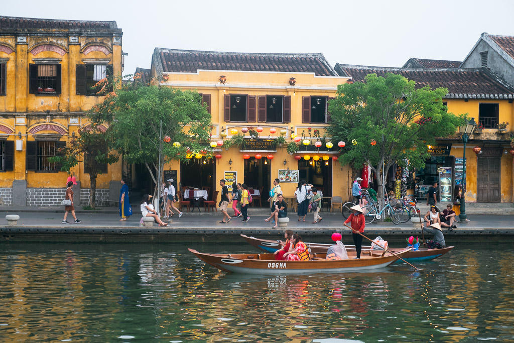 People walking on a sidewalk in front of yellow two-story buildings and people riding a boat on a river in Hoi An, Vietnam.