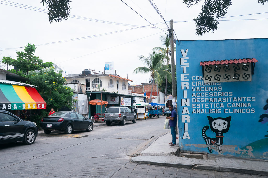 Street scene in Puerto Escondido, Mexico, featuring a vibrant blue building, a colorful striped awning, and several parked cars lining the road under tropical trees.