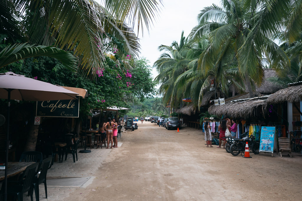 Dusty palm-lined street in La Punta, Oaxaca, with small cafes, thatched-roof shops, and people walking by