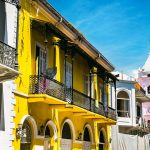 A yellow building with ornate balconies, set against a backdrop featuring a historic bell tower, in Casco Viejo, Panama.