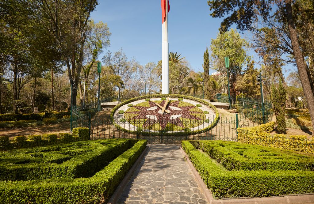A park in Mexico City with a flag pole and a landscape art that looks like a giant clock.