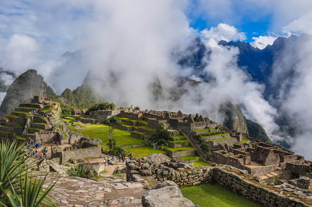 View of Machu Picchu’s ancient stone ruins surrounded by misty clouds and lush green terraces, with Andean mountains rising in the background