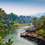 The River Kwai in Thailand surrounded by houses, trees, railroad, and mountains in the background.