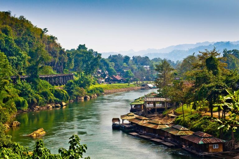 The River Kwai in Thailand surrounded by houses, trees, railroad, and mountains in the background.