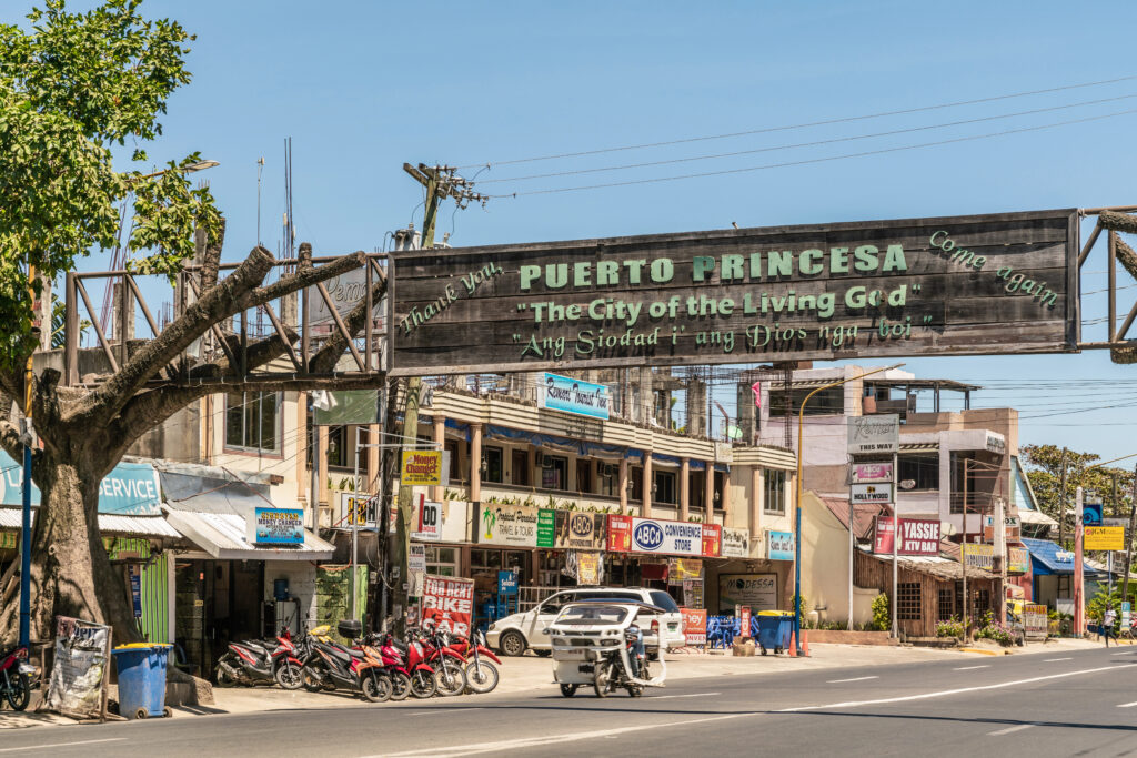 Street scene in Puerto Princesa, Palawan, Philippines, featuring a wooden sign that reads "Puerto Princesa 'The City of the Living God'"