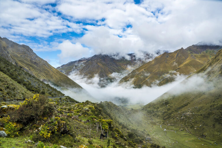 A hazy, high-altitude view of the Salkantay Trail, with mist filling the valleys between steep, grassy mountains.