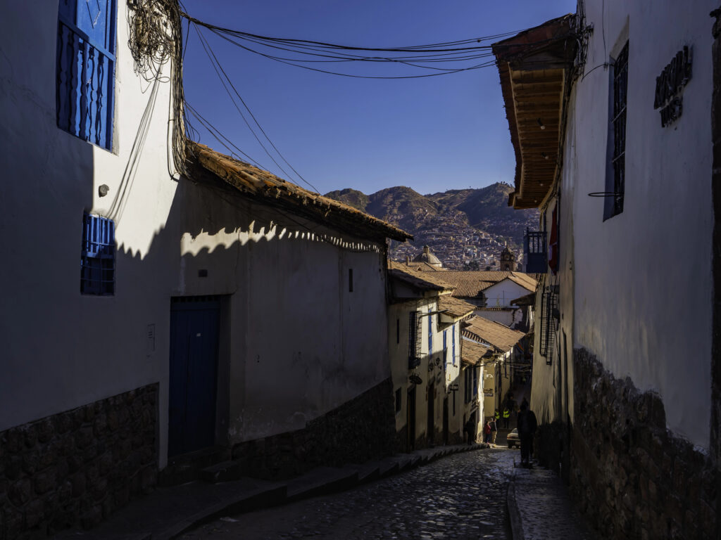 Sunlit street between white buildings with blue accents, leading to distant mountains in old quarter Cusco, Peru