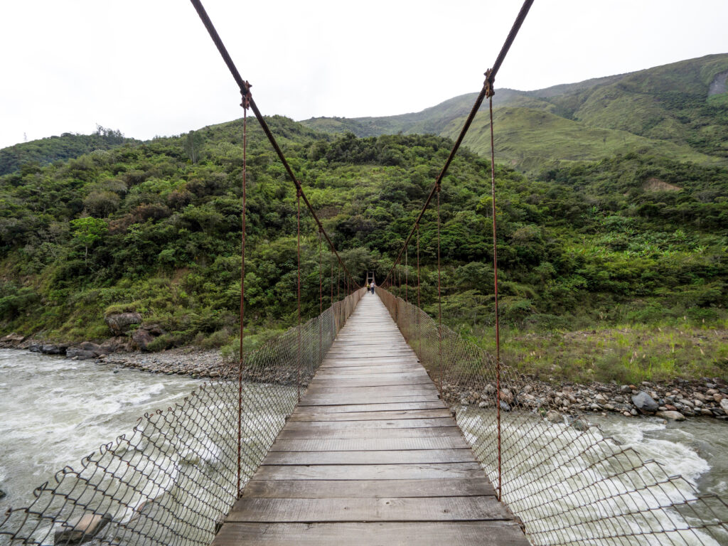 A long, wooden cable suspension bridge stretches across a rushing, rocky river, likely the Urubamba River, in a lush, mountainous region of the Inca Jungle Trek.
